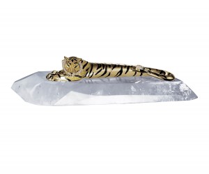 Stylo Plume D’Exception Décor Tigre – Maison Cartier « Telle une sculpture animalière, ce stylo plume à décor tigre, héros du bestiaire précieux Cartier, est ici saisi dans toute la plénitude de son repos. Animal d’Afrique, nouvelle terre d’inspiration pour Cartier, qui réalise grâce à un moulage précis, la silhouette du félin allongé sur un socle en cristal de roche, véritable savoir-faire issu de la joaillerie. Plus de 100 heures de travail ont été nécessaire à la création de ce stylo tout en contraste chromatique. Le seul sertissage des centaines de pierres qui composent ce stylo de prestige requiert plus d’une dizaine d’heures. La technique de laquage pour la réalisation des tigrures demande un soin très particulier : la laque est déposée à la seringue, tigrure par tigrure, puis patiemment polie pour obtenir un résultat parfait. La plume en or massif est gravée d’une tête de tigre. Le socle est réalisé en cristal de roche, sculpté à même un bloc de cristal. Cette opération, très délicate, demande un savoir-faire rare, privilège de seulement quelques ateliers dans le monde. Avec ce stylo plume d’exception, l’écriture entre dans le décor et signe sa présence joaillière au fil de détails d’excellence : tête pavée de diamants blancs et jaunes, yeux en émeraude, truffe en onyx, tâches en laque noire, plume en or massif… Un objet précieux en édition limitée à 5 exemplaires dont l’esthétique et l’énergie des lignes traduisent la puissance du félin et l’art de Cartier, joaillier de l’animalier. »
