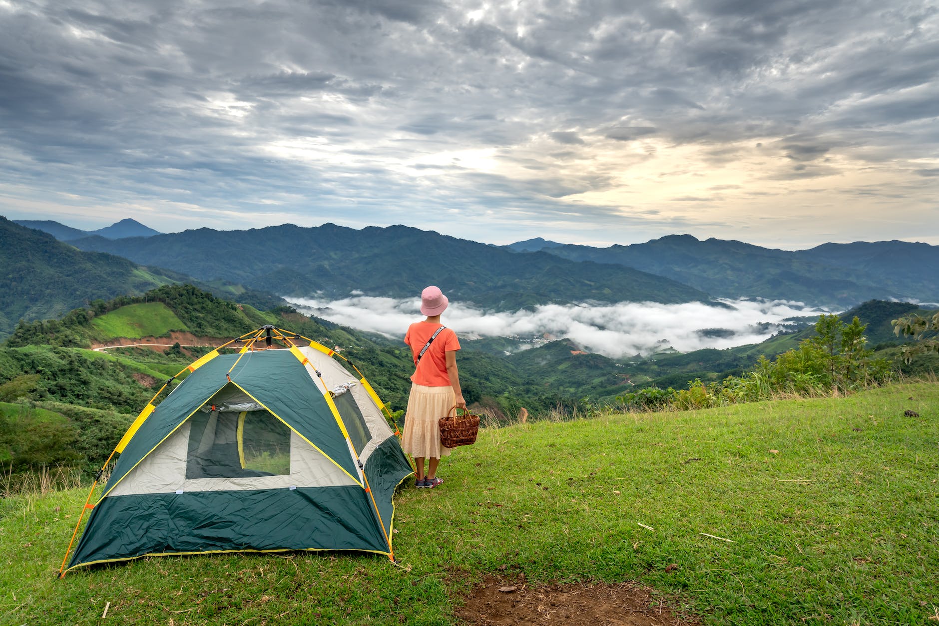 a woman in orange shirt standing beside a tent on a green grass field while looking at the foggy mountains