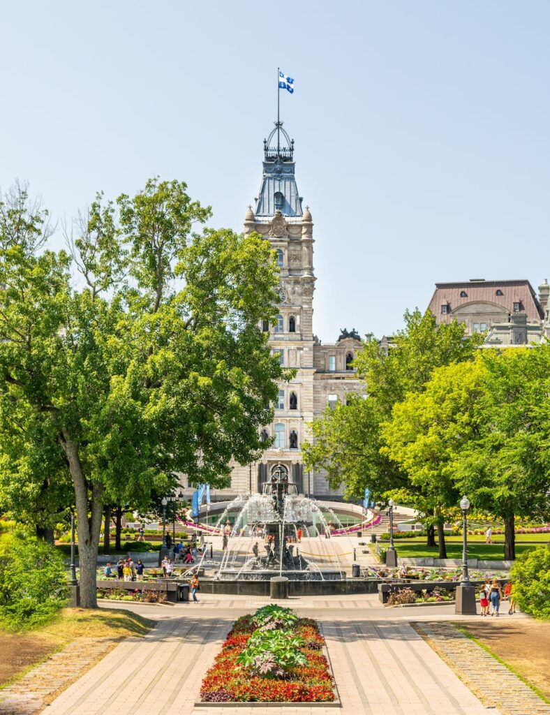 people by a fountain in the park with a walkway surrounded with trees and flowers with in front of a building with a tower